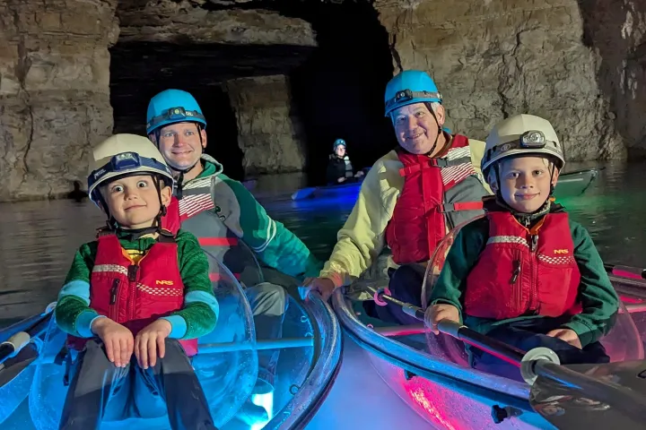 People in helmets kayaking in a dimly lit cave with illuminated kayaks.