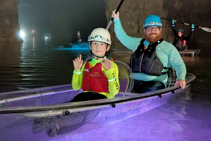 Two people kayak in a clear boat inside a dimly lit cave.